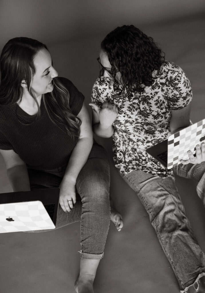 two virtual assistant sitting on the floor together working on their laptops together while laughing